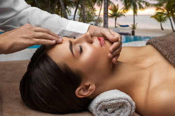 Fototapeta na wymiar wellness, beauty and relaxation concept - beautiful young woman lying with closed eyes and having face and head massage at spa over tropical beach background in french polynesia