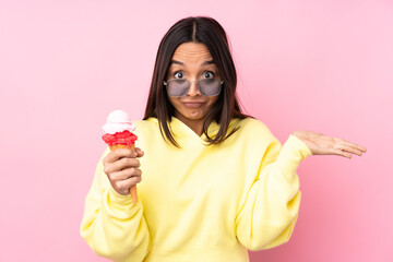 Young brunette girl holding a cornet ice cream over isolated pink background having doubts while...