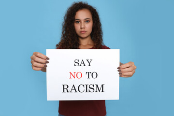 African American woman holding sign with phrase Say No To Racism against light blue background,...