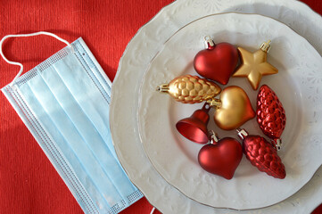 Christmas table. white plates with toys, a knife and fork on a red background and a medical protective mask. COVID. Preparation for Christmas and New Year.