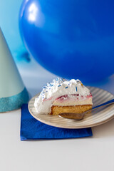 food, celebration and holidays concept - piece of birthday cake on plate with spoon, party hat and blue balloons on table