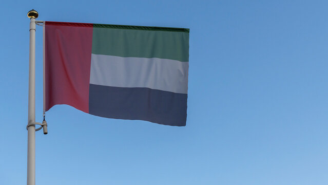 National flag of United Arab Emirates on a flagpole in front of blue sky with sun rays and lens flare.