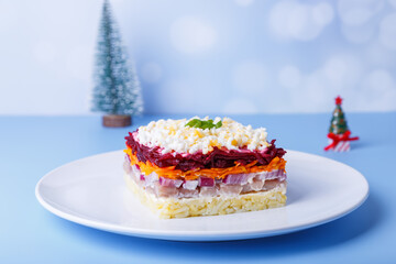 Herring salad under a fur coat. Traditional Russian multilayered salad from herring, beets, potatoes, carrots and eggs. Close-up, blue background, miniature Christmas tree.
