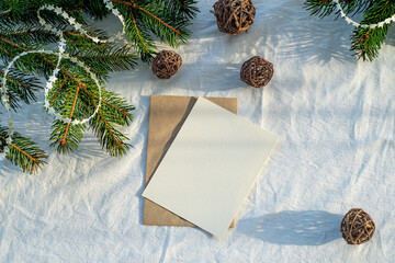 Blank greeting card, invitation card mockup.Christmas decoration scene in sunlight.Card envelope, ribbon, fir branches and wooden balls.White linen background.Top view, flat lay.Winter wedding concept