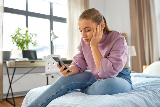 children, technology and communication concept - sad teenage girl with smartphone sitting on bed at home