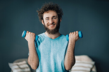 Handsome Man Does Fitness with Dumbbells At Home. Guy Keeps Fit During Quarantine. Gray background - obrazy, fototapety, plakaty