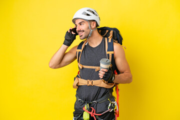 Fototapeta premium Young caucasian rock climber man isolated on yellow background holding coffee to take away and a mobile