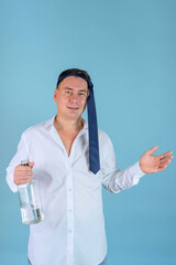 A drunk businessman with a tie on his head and a bottle of alcohol in his hands laughs and spreads his arms standing on a blue background. Vertical photo