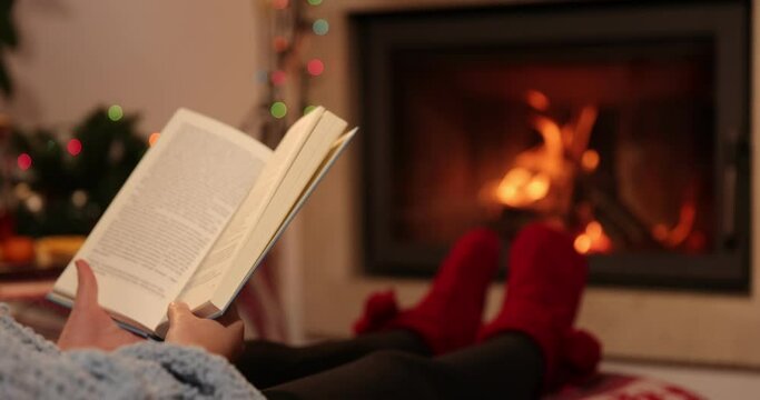 Girl Reading a Book by the Fireplace. Slow motion 4K. Young woman reading a book by the warm fireplace decorated for Christmas. Relaxed holiday evening concept
