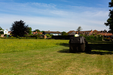 A council worker riding on a sit on lawnmower cutting the grass in a public park on a hot summers day