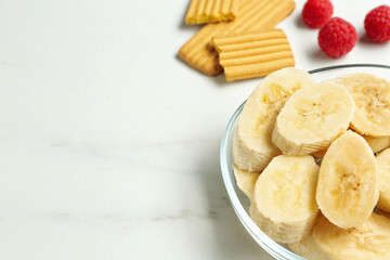 Cut bananas in glass bowl and other baby finger foods on white marble table, space for text