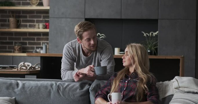 European blonde couple talking relaxing together at comfortable loft apartment interior. Handsome boyfriend and beautiful boyfriend drinking tea coffee holding mug discussing news planning dialogue