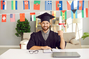 Portrait of happy guy in grad mortarboard hat and graduate gown sitting at desk, holding...