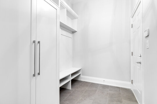 A Bright, White Mud Room With A Closet, Shelving, A Bench For Seating And A Dark Tiled Floor.