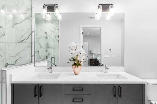 A beautiful bathroom with a dark vanity and white granite counter top. A plant and towels sit on the vanity. The shower is lined with marble tiles. Lights on.