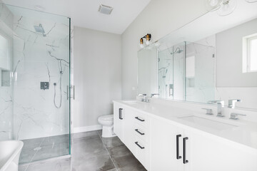 A modern, white luxury bathroom with black hardware and chrome faucets. A large glass walled shower sits in the corner with marble tiles from floor to ceiling. - obrazy, fototapety, plakaty