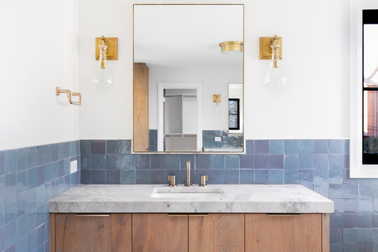A luxury home's renovated bathroom with blue tiles, a light wood vanity cabinet, gold lights mounted on the wall, and a grey stone counter top.