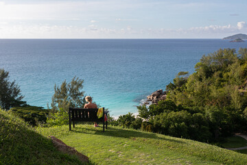 Fototapeta premium A young woman is sitting on a black wooden bench on a high green hill. The hill is opposite the blue endless ocean. Coast consist of huge granite rocks, white sand beach and green tropical forest.