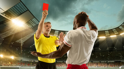 Football referee showing a player a red card for breaking rules at crowded stadium over night cloudy sky.
