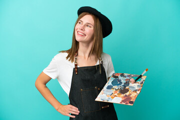 Young artist English woman holding a palette isolated on blue background posing with arms at hip and smiling