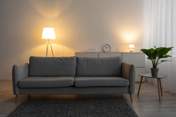 Couch, clock, plant in pot on table and glowing lamp in evening