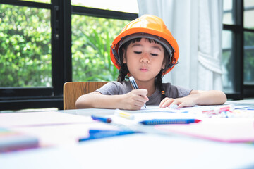 Blurred soft images of Asian girl, 6-year-old wearing engineer helmet, is coloring with intention,...
