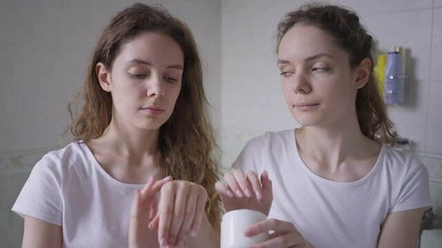 Funny young woman touching twin sister nose with moisturizer laughing as siblings applying hand cream in bathroom at home in the morning. Cheerful Caucasian women having fun taking care of skin
