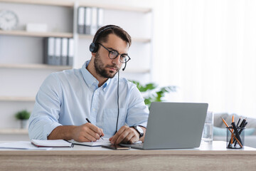 Busy adult european man manager with beard in glasses and headphones work at laptop in office