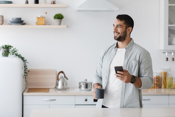 Smiling middle aged caucasian male with beard with smartphone reading news and drinking coffee
