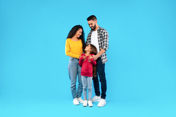 Middle-Eastern Parents Embracing Their Happy Daughter On Blue Background