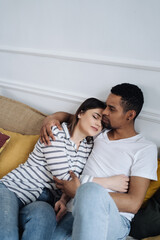 Obraz premium Portrait of happy young African Caucasian couple hug sitting on couch at home, smiling mixed race husband and wife spend time together, embrace relaxing on sofa. Healthy relationships concept