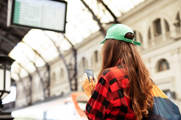 Young woman waiting for the train standing at the railway station. Beautiful girl typing a message while waiting for the train.