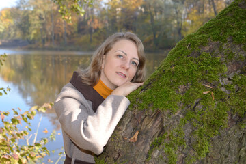 The woman leaned against an old mossy tree against the backdrop of the lake. Autumn Park