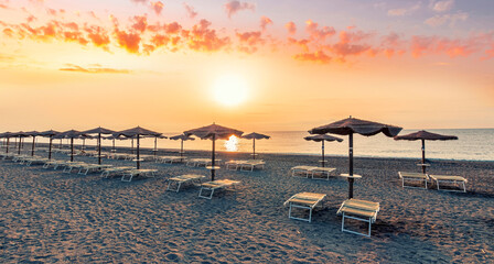 scenic view at nice sunset or sunrise beach with rows of umbrellas and chaise lounges with blue sea and amazing cloudy sky with sun glow on the background