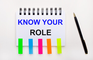 On a light background, a pen, a notebook with the text KNOW YOUR ROLE and bright stickers