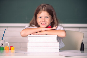 Smiling girl sitting at the desk and holding hands on the books in the school classroom. Educational process.