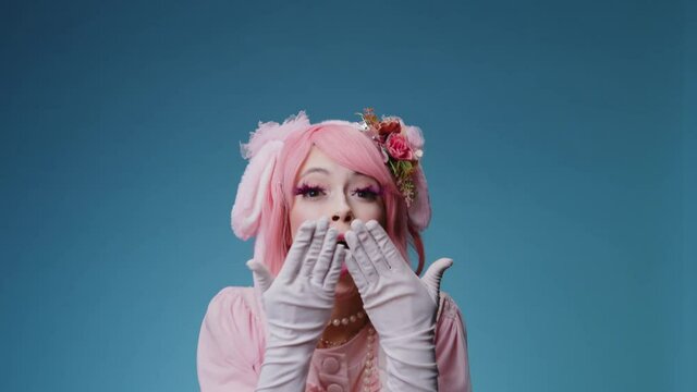 Shy and playful glamour young sexy candy girl with pink makeup and fancy clothes sends an air kiss on blue background. Millenial generation fashion girl flirting concept