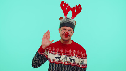 Man in Christmas sweater, deer antlers waves hand palm in hello gesture welcomes someone with hospitable expression expresses positive emotions on blue studio background. Happy New Year celebration