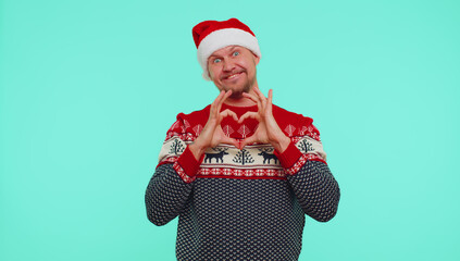 Smiling man in Christmas sweater makes heart gesture demonstrates love sign expresses good feelings and sympathy. Young adult guy isolated on blue studio background. Happy New Year celebration holiday