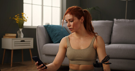 Beautiful young woman in sportswear resting after intensive home workout. Athletic lady finished fitness training, sitting tired on floor holding sport water bottle and mobile phone.