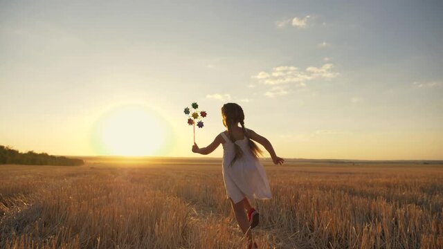 little girl runs across field at sunset with windmill in her hands. Silhouette of child girl holding wind toy. child enjoys fresh air. fun game in wind in field. girl runs at sunset.