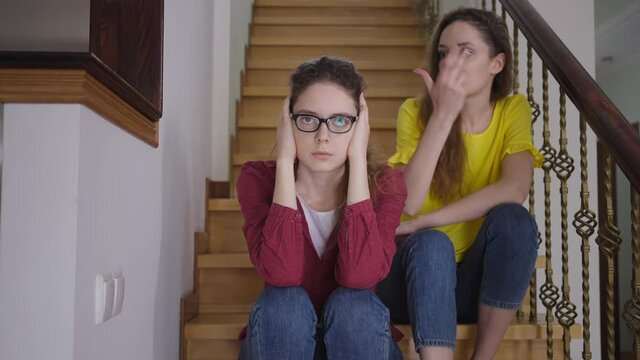 Tired woman in eyeglasses covering ears with hands sighing as excited twin sister talking sitting on stairs at background. Portrait of skeptic intelligent Caucasian nerd with cheerful sibling at home