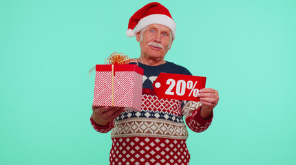 Senior grandfather in New Year sweater showing gift box and 20 Percent discount inscriptions banner text. Black Friday. Holiday low prices for shopping. Blue studio wall background. Merry Christmas
