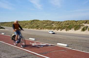 Red bike path in the Netherlands. Route on a dam in front of the grass dunes. Man on bicycle motionless. Car driving on the road. Schouwen-Duiveland, Brouwersdam.