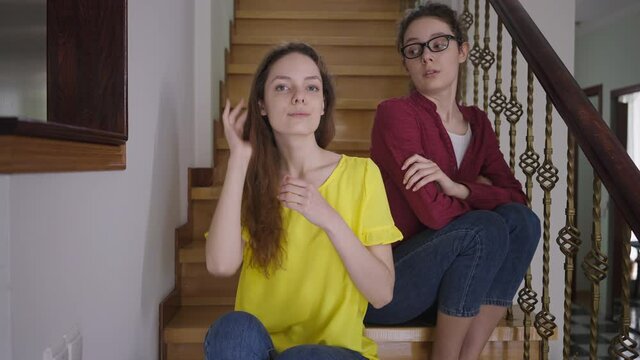 Portrait of confident slim beautiful woman looking at camera touching hair with sarcastic twin sister in eyeglasses sitting at background on stairs. Narcissistic Caucasian lady with nerd sibling