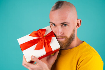 A bearded man in an orange T-shirt on a turquoise background with a gift box in an elated mood, gives joy, smiles pleasantly