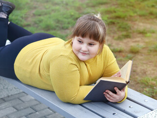 Plump smiling girl lying on bench in park, holds a book in her hands and looks away