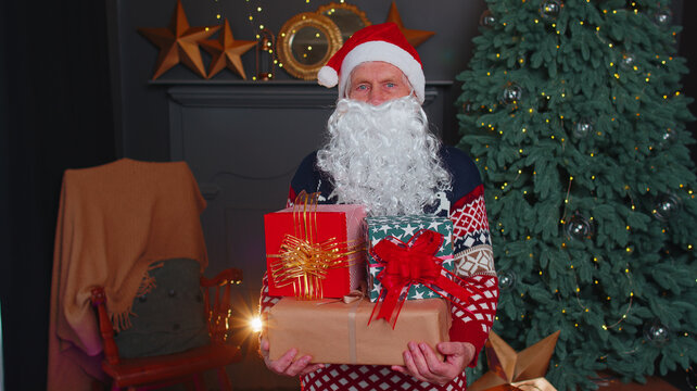 Mature Grandfather Wears Red Festive Sweater, Fake Beard Presenting Christmas Gift Boxes In Cozy Living Room At Home. Happy Mature Man Parodies Santa Claus On New Year Xmas Celebration Holidays Eve