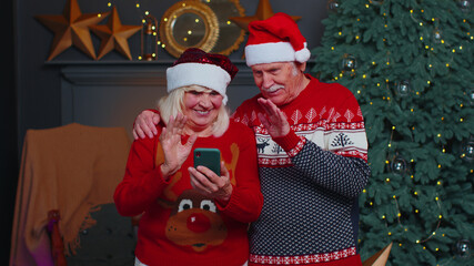 Happy elderly family couple grandmother grandmother standing at decorated home room with Christmas tree taking selfie on mobile phone, communicating video call online celebrating New Year together