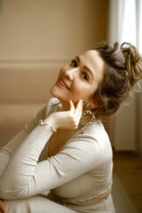 portrait of young caucasian woman in a beige house suit sitting on the floor, a beautiful lady with large earrings and high hairstyle in a stylish apartment, a stylish image of a young successful